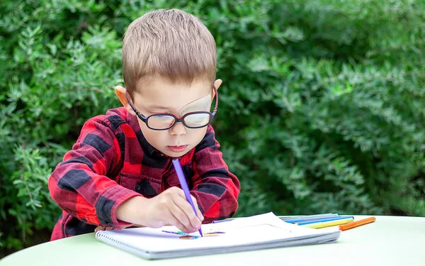 Ein Junge mit Amblyopie hat sein linkes Auge mit einem Okkluder abgeklept, er trägt dazu eine Brille und sitzt malend im Garten an einem Tisch.