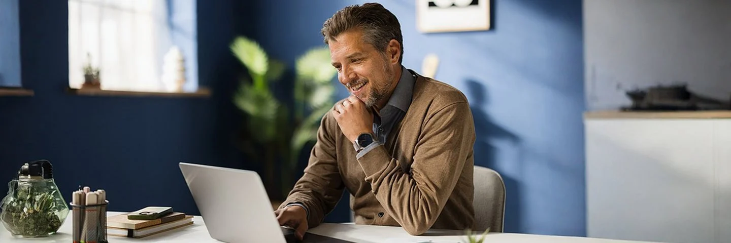 Ein Mann mit einer braunen Strickjacke und grauem Hemd sitzt lächelnd in einem blauen Arbeitszimmer vor einem aufgeklappten Laptop.
