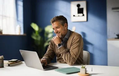 Ein Mann mit einer braunen Strickjacke und grauem Hemd sitzt lächelnd in einem blauen Arbeitszimmer vor einem aufgeklappten Laptop.