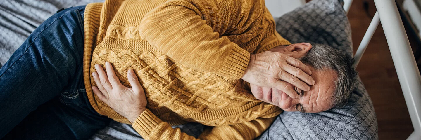 Ein Mann mit Darmverschluss liegt auf dem Bett; eine Hand hält er vor seinem Gesicht, die andere auf seinem schmerzenden Unterbauch.