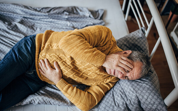 Ein Mann mit Darmverschluss liegt auf dem Bett; eine Hand hält er vor seinem Gesicht, die andere auf seinem schmerzenden Unterbauch.