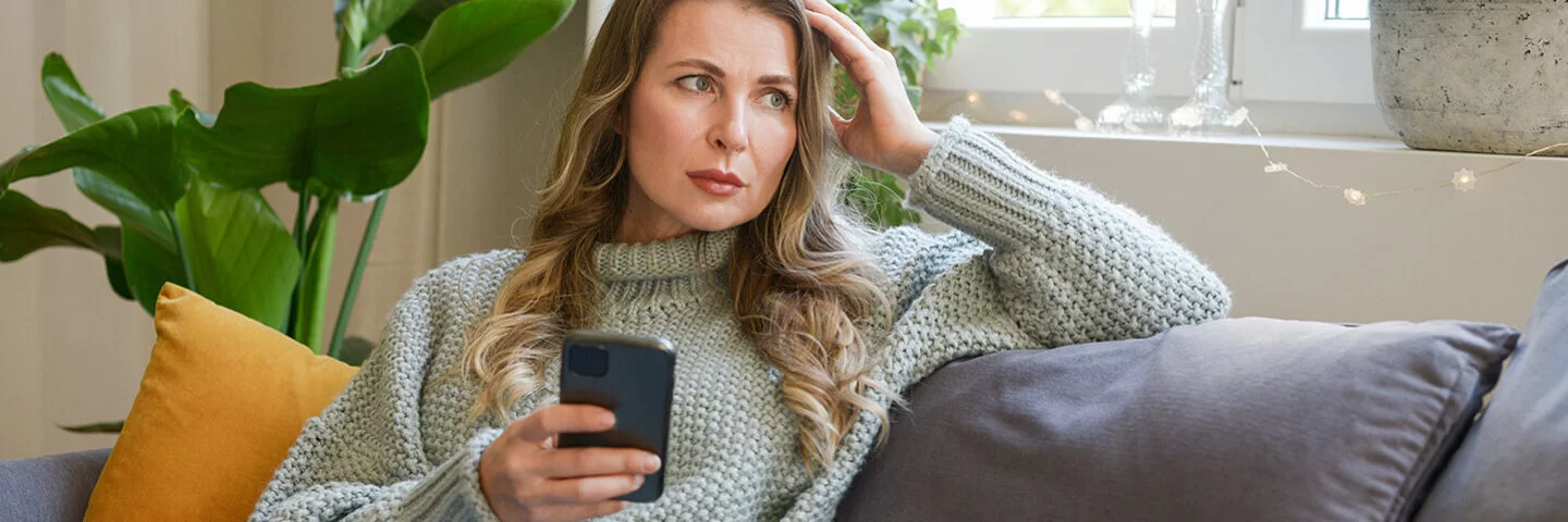 Eine Frau mittleren Alters sitzt in einer Wohnung auf dem Sofa. In der rechten Hand hält sie ein Smartphone, mit der linken stützt sie ihren Kopf. Sie blickt ins Leere. Ihre Mimik vermittelt Enttäuschung.