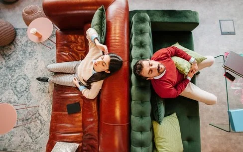 Zwei Personen liegen auf gegenüberstehenden Sofas, eine auf einem braunen Ledersofa mit einem Buch, die andere auf einem grünen Samtsofa mit einem Kissen.