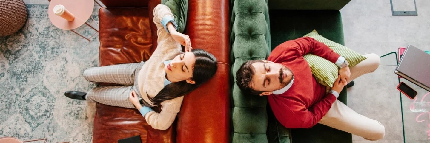 Zwei Personen liegen auf gegenüberstehenden Sofas, eine auf einem braunen Ledersofa mit einem Buch, die andere auf einem grünen Samtsofa mit einem Kissen.