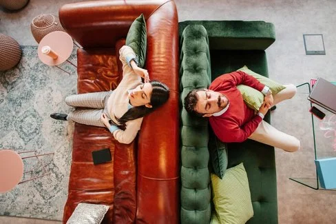 Zwei Personen liegen auf gegenüberstehenden Sofas, eine auf einem braunen Ledersofa mit einem Buch, die andere auf einem grünen Samtsofa mit einem Kissen.