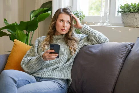 Eine Frau mittleren Alters sitzt in einer Wohnung auf dem Sofa. In der rechten Hand hält sie ein Smartphone, mit der linken stützt sie ihren Kopf. Sie blickt ins Leere. Ihre Mimik vermittelt Enttäuschung.