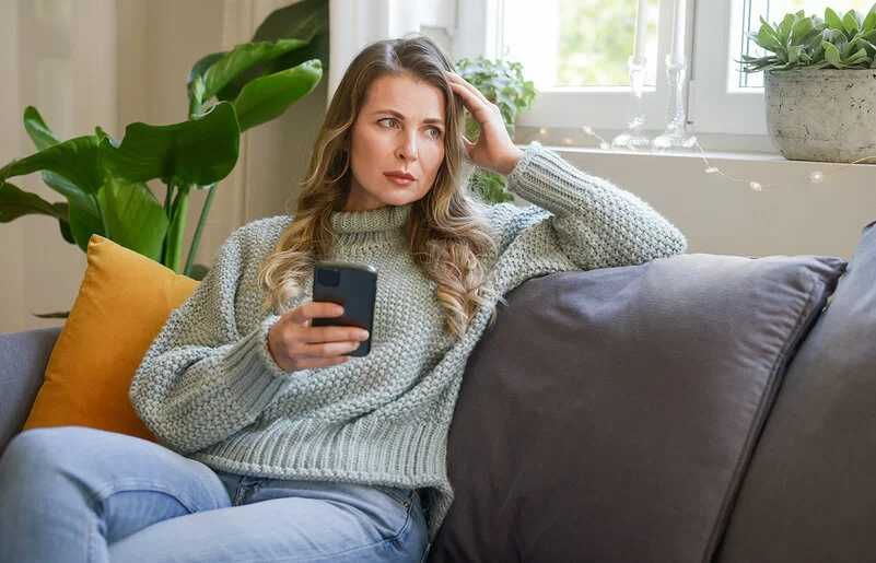 Eine Frau mittleren Alters sitzt in einer Wohnung auf dem Sofa. In der rechten Hand hält sie ein Smartphone, mit der linken stützt sie ihren Kopf. Sie blickt ins Leere. Ihre Mimik vermittelt Enttäuschung.