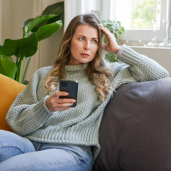 Eine Frau mittleren Alters sitzt in einer Wohnung auf dem Sofa. In der rechten Hand hält sie ein Smartphone, mit der linken stützt sie ihren Kopf. Sie blickt ins Leere. Ihre Mimik vermittelt Enttäuschung.