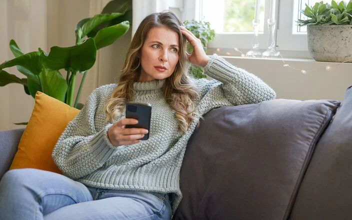Eine Frau mittleren Alters sitzt in einer Wohnung auf dem Sofa. In der rechten Hand hält sie ein Smartphone, mit der linken stützt sie ihren Kopf. Sie blickt ins Leere. Ihre Mimik vermittelt Enttäuschung.