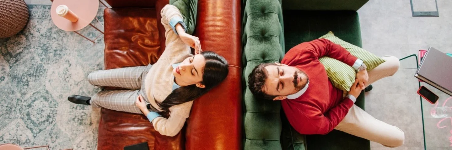 Zwei Personen liegen auf gegenüberstehenden Sofas, eine auf einem braunen Ledersofa mit einem Buch, die andere auf einem grünen Samtsofa mit einem Kissen.
