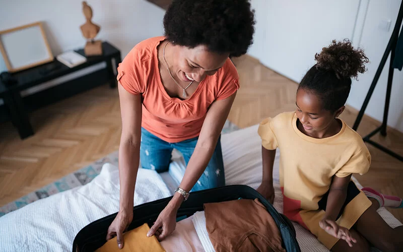 Mutter und Tochter packen fröhlich gemeinsam den Koffer des Kindes im Schlafzimmer.