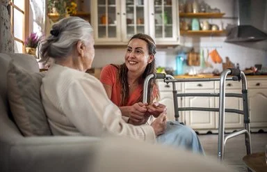 Eine Frau mit dunkeln Haaren und orangen T-Shirt hockt neben einer älteren Frau, die im Sessel sitzt. Sie sind sich freundlich zugewandt. Neben ihnen steht ein Rollator. Im Hintergrund sind ein Wohnbereich und eine Küche zu sehen.