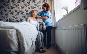 Eine Pflegerin hilft einer älteren Frau beim Ankleiden. Die ältere Frau sitz in ihrem Zuhause auf dem Bett. Sie bekommt eine Bluse angezogen.