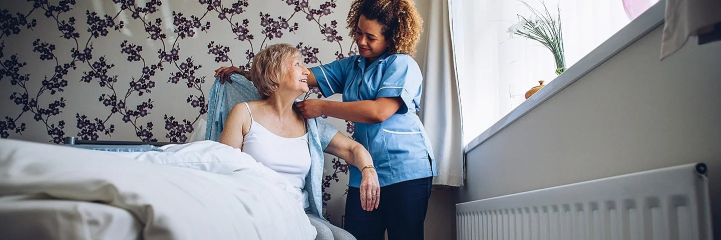 Eine Pflegerin hilft einer älteren Frau beim Ankleiden. Die ältere Frau sitz in ihrem Zuhause auf dem Bett. Sie bekommt eine Bluse angezogen.