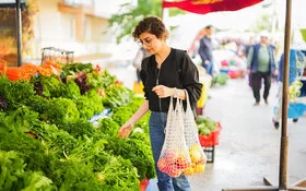 Eine Frau steht vor einem Gemüsestand auf dem Wochenmarkt und betrachtet die ausliegenden Blattsalate. An ihrem linken Arm hängen zwei Einkaufsnetze mit Obst.