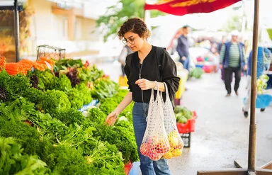 Eine Frau steht vor einem Gemüsestand auf dem Wochenmarkt und betrachtet die ausliegenden Blattsalate. An ihrem linken Arm hängen zwei Einkaufsnetze mit Obst.