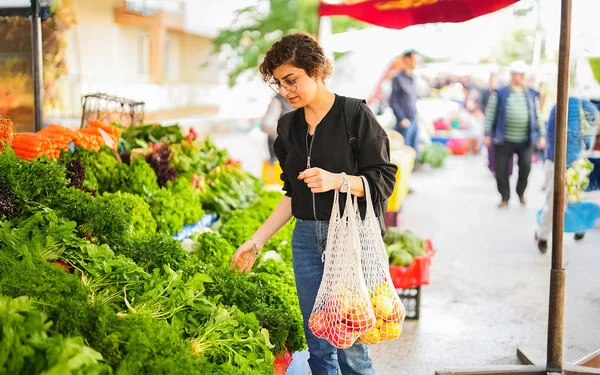 Eine Frau steht vor einem Gemüsestand auf dem Wochenmarkt und betrachtet die ausliegenden Blattsalate. An ihrem linken Arm hängen zwei Einkaufsnetze mit Obst.