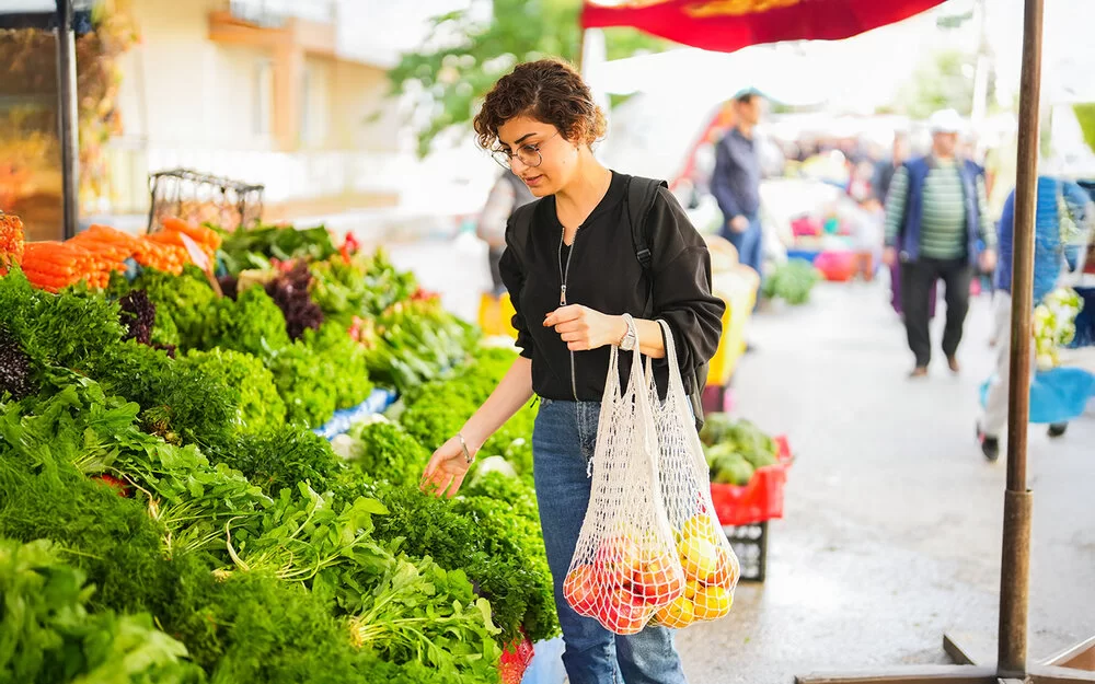 Eine Frau steht vor einem Gemüsestand auf dem Wochenmarkt und betrachtet die ausliegenden Blattsalate. An ihrem linken Arm hängen zwei Einkaufsnetze mit Obst.