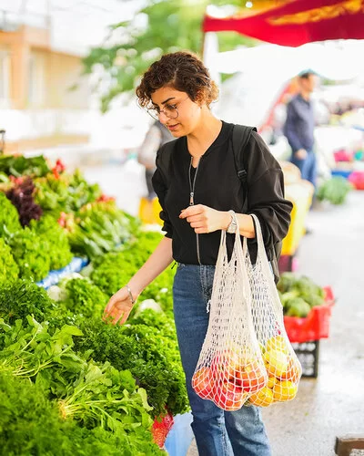 Eine Frau steht vor einem Gemüsestand auf dem Wochenmarkt und betrachtet die ausliegenden Blattsalate. An ihrem linken Arm hängen zwei Einkaufsnetze mit Obst.