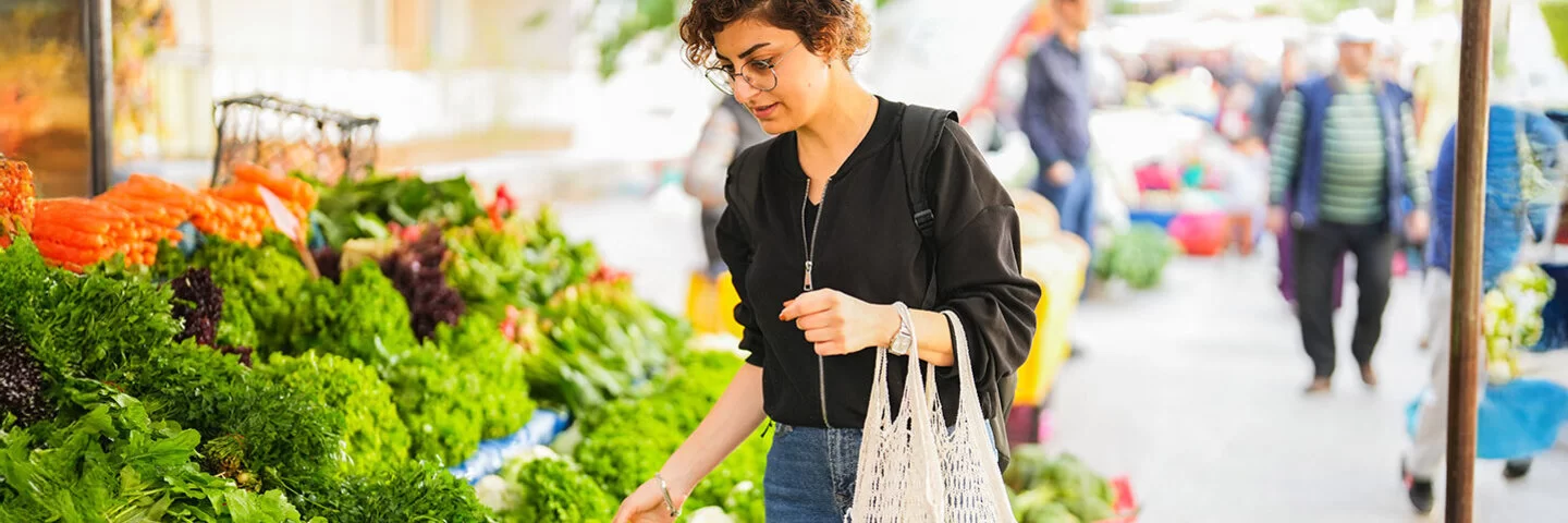 Eine Frau steht vor einem Gemüsestand auf dem Wochenmarkt und betrachtet die ausliegenden Blattsalate. An ihrem linken Arm hängen zwei Einkaufsnetze mit Obst.