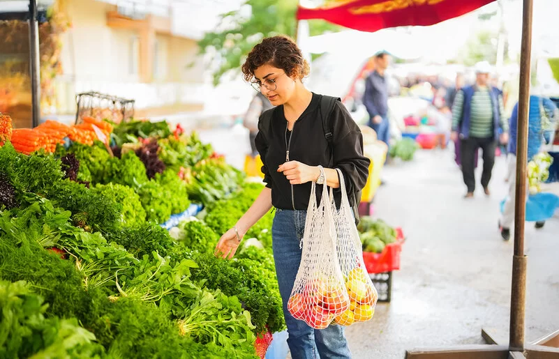 Eine Frau steht vor einem Gemüsestand auf dem Wochenmarkt und betrachtet die ausliegenden Blattsalate. An ihrem linken Arm hängen zwei Einkaufsnetze mit Obst.