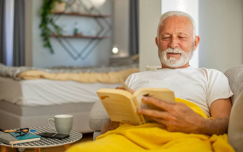 Ein älterer Mann liegt auf der Couch und liest mit leicht zusammengekniffenen Augen ein Buch.