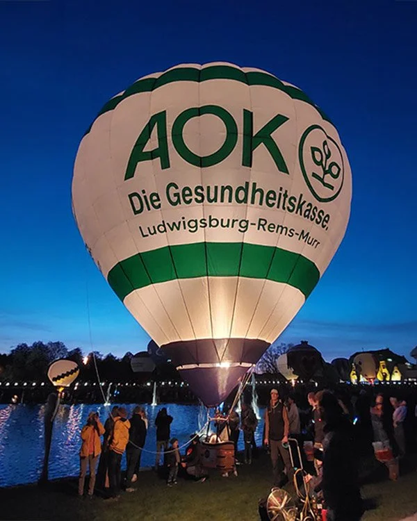 Heißluftballon mit der Aufschrift 'AOK Die Gesundheitskasse Ludwigsburg-Rems-Murr' bei Dämmerung am Wasser mit Menschen im Vordergrund.