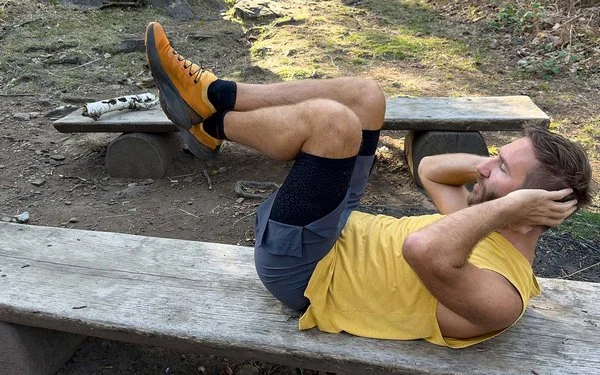 Ein Mann in Sportkleidung liegt auf einer langen Holzbank im Wald und macht Bauchpressen.