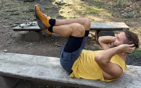 Ein Mann in Sportkleidung liegt auf einer langen Holzbank im Wald und macht Bauchpressen.