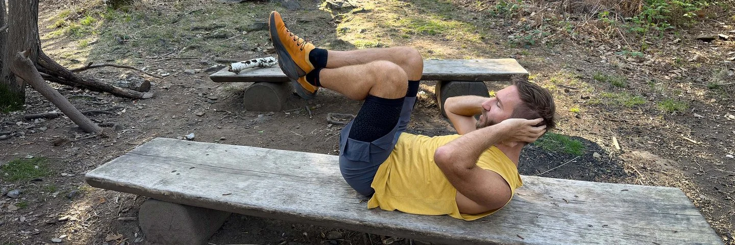 Ein Mann in Sportkleidung liegt auf einer langen Holzbank im Wald und macht Bauchpressen.