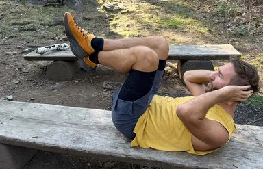 Ein Mann in Sportkleidung liegt auf einer langen Holzbank im Wald und macht Bauchpressen.