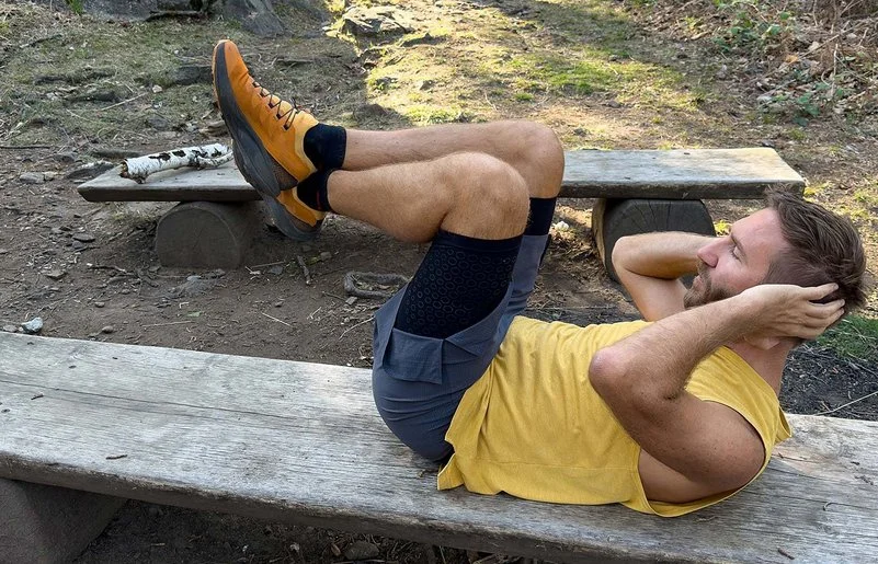 Ein Mann in Sportkleidung liegt auf einer langen Holzbank im Wald und macht Bauchpressen.