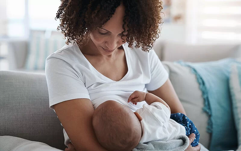 Eine junge Mutter im weißen T-Shirt sitzt auf einem Sofa, hält ihr Baby im Arm und stillt es. Dabei schaut sie es liebevoll an.