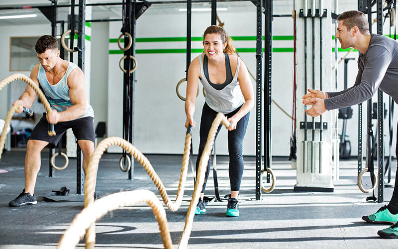 In einem Fitnessstudio absolvieren ein Mann und eine Frau ein Battle-Rope-Workout mit einem Trainer.