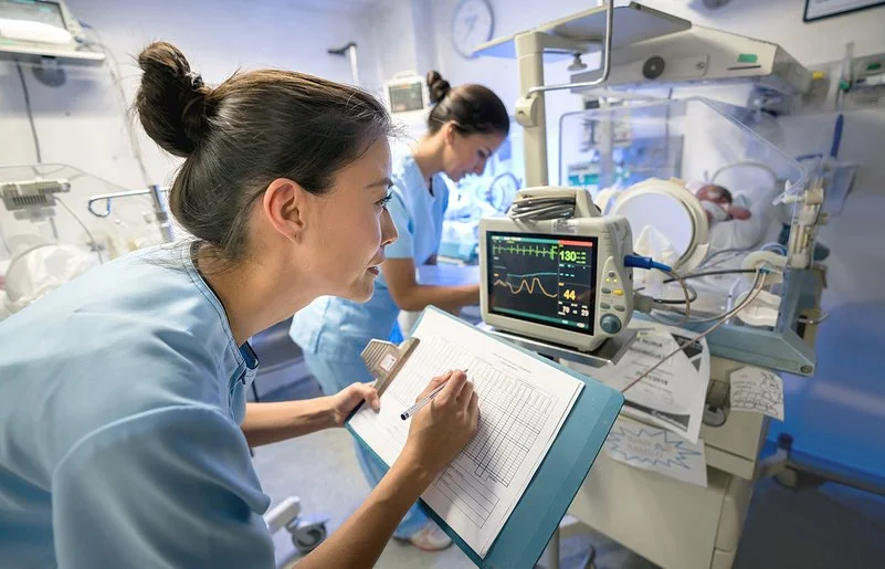 Intensivpflege: Zwei Pflegefachpersonen versorgen ein Baby, das in einem Brutkasten liegt.