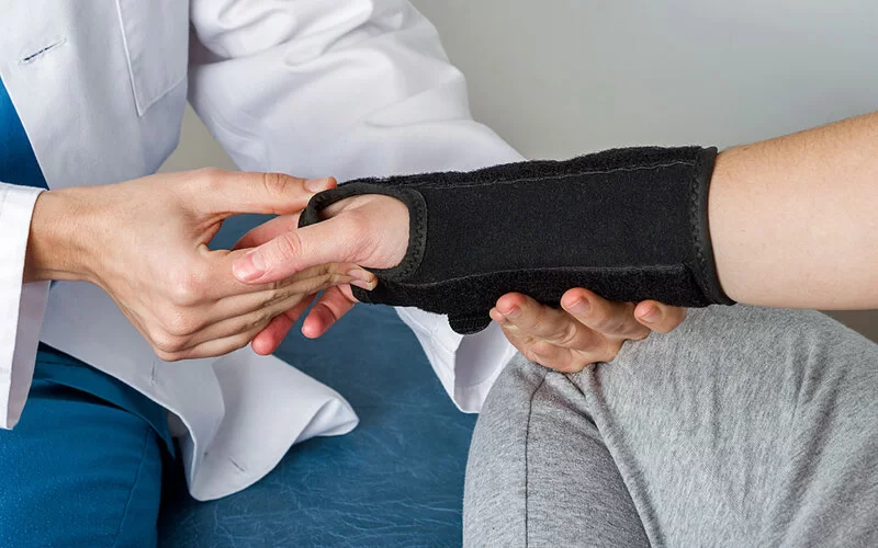 Ein Arzt untersucht die Hand einer Patientin, die eine schwarze Orthese trägt.