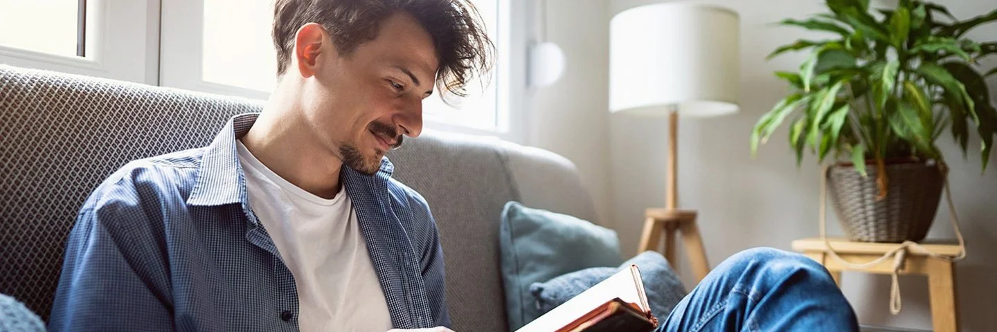 Junger Mann mit dunklen Haaren sitzt entspannt auf einem Sofa am Fenster und liest ein Buch. Er trägt eine blaue Jeans, ein offenes blaues Hemd und ein weißes T-Shirt. Im Hintergrund ist ein helles, gemütliches Wohnzimmer mit Zimmerpflanze und Stehlampe zu sehen.