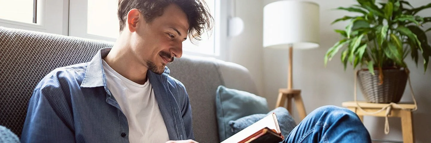 Junger Mann mit dunklen Haaren sitzt entspannt auf einem Sofa am Fenster und liest ein Buch. Er trägt eine blaue Jeans, ein offenes blaues Hemd und ein weißes T-Shirt. Im Hintergrund ist ein helles, gemütliches Wohnzimmer mit Zimmerpflanze und Stehlampe zu sehen.
