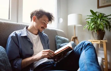 Junger Mann mit dunklen Haaren sitzt entspannt auf einem Sofa am Fenster und liest ein Buch. Er trägt eine blaue Jeans, ein offenes blaues Hemd und ein weißes T-Shirt. Im Hintergrund ist ein helles, gemütliches Wohnzimmer mit Zimmerpflanze und Stehlampe zu sehen.