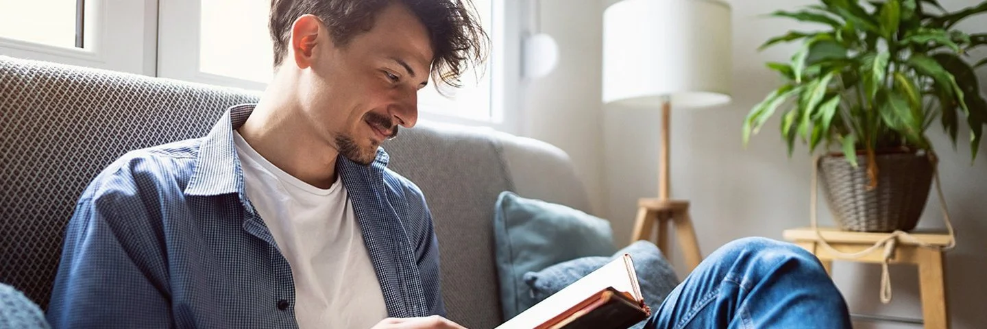 Junger Mann mit dunklen Haaren sitzt entspannt auf einem Sofa am Fenster und liest ein Buch. Er trägt eine blaue Jeans, ein offenes blaues Hemd und ein weißes T-Shirt. Im Hintergrund ist ein helles, gemütliches Wohnzimmer mit Zimmerpflanze und Stehlampe zu sehen.