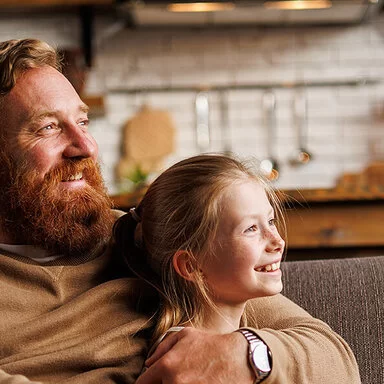Bärtiger Mann lächelt und umarmt vorpubertäre Tochter, während er sich zu Hause auf der Couch entspannt. Familien sind bei der AOK gut versichert.
