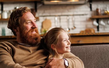 Bärtiger Mann lächelt und umarmt vorpubertäre Tochter, während er sich zu Hause auf der Couch entspannt. Familien sind bei der AOK gut versichert.