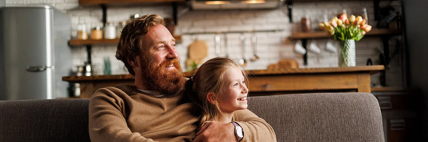 Bärtiger Mann lächelt und umarmt vorpubertäre Tochter, während er sich zu Hause auf der Couch entspannt. Familien sind bei der AOK gut versichert.
