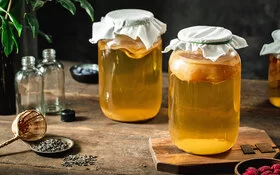 Zwei mit Tüchern abgedeckte Gläser mit Kombucha-Teegetränk und SCOBY auf einem Tisch.