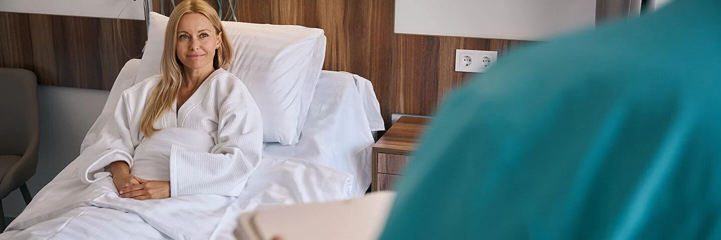 Ein Frau sitzt aufrecht in einem Krankenbett, ihre Hände liegen gefaltet auf ihrer Bettdecke. Im Vordergrund ist eine Pflegeperson in einem blaugruenen Arbeitskittel zu sehen.