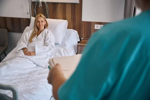 Ein Frau sitzt aufrecht in einem Krankenbett, ihre Hände liegen gefaltet auf ihrer Bettdecke. Im Vordergrund ist eine Pflegeperson in einem blaugruenen Arbeitskittel zu sehen.