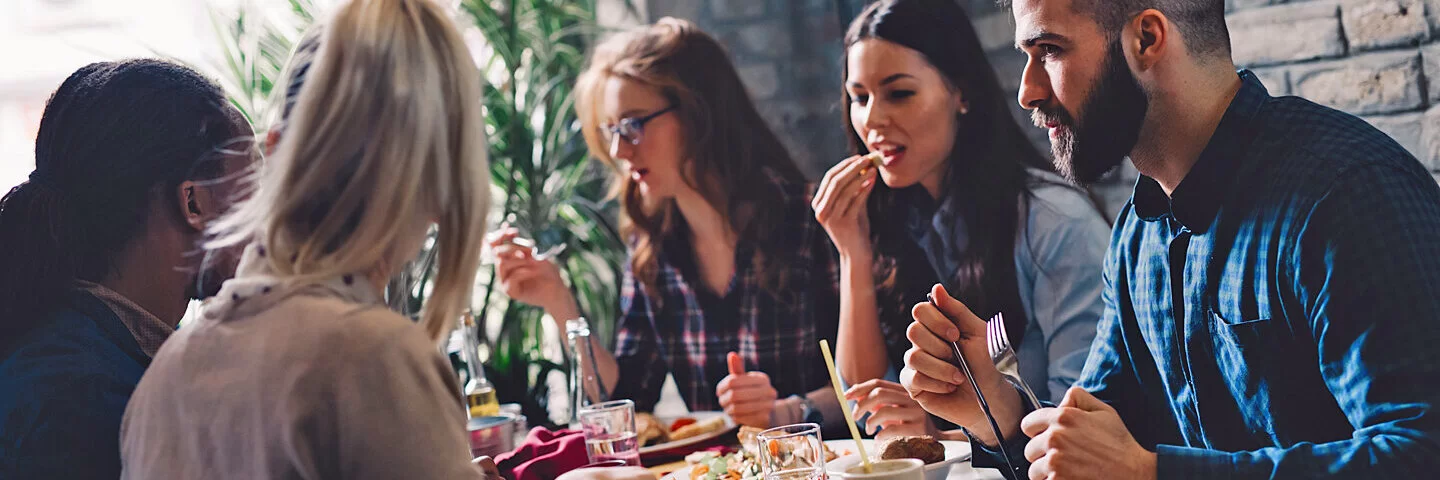 Ein Gruppe junger Menschen bei einem Geschäftsessen, eine von ihnen fürchtet, die anderen könnten ihre lauten Darmgeräusche hören.