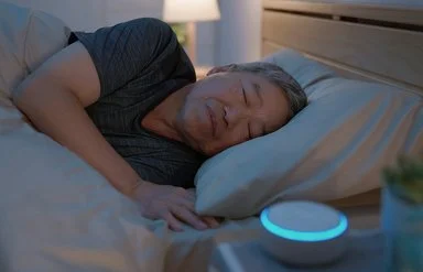 Ein Mann liegt im Bett und schläft. Auf dem Nachttisch neben ihm steht eine kleine, kreisrunde „White-Noise-Machine”.
