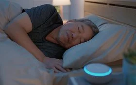 Ein Mann liegt im Bett und schläft. Auf dem Nachttisch neben ihm steht eine kleine, kreisrunde „White-Noise-Machine”.