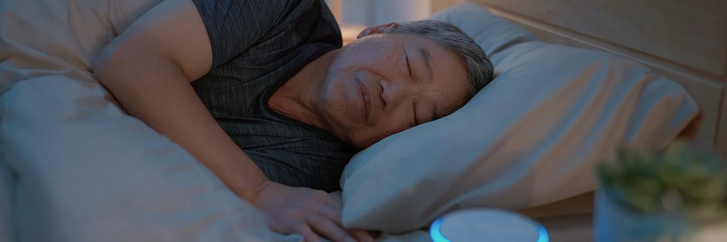 Ein Mann liegt im Bett und schläft. Auf dem Nachttisch neben ihm steht eine kleine, kreisrunde „White-Noise-Machine”.